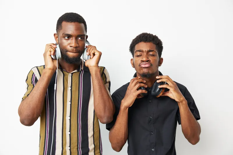 Two black men enjoying music together with headphones, conveying a fun and relaxed atmosphere while showcasing their unique sense of style and rhythm.