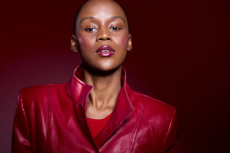 Portrait of confident African woman with dark skin wearing glossy red leather jacket and matching top, posing against deep red background. Concept of fashion, beauty, and elegance.