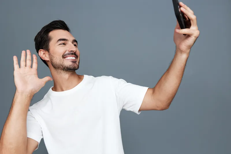 Happy smiling young man waving at the camera while taking a selfie with his smartphone, dressed in a casual white t-shirt against a gray background.