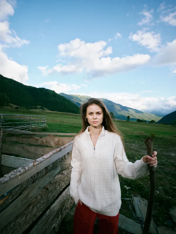 Farmer woman stands in an open field with a pitchfork, capturing rural landscape and outdoor life, casual sweater, wooden fence, expansive countryside