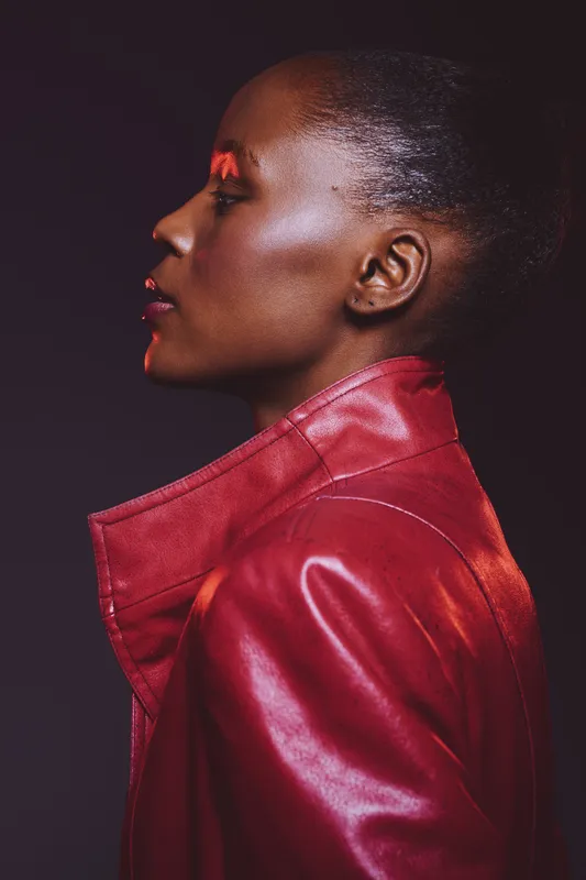 Profile portrait of an elegant dark skinned African woman wearing a red leather jacket against a dark background. Concept of fashion, beauty, confidence, and style.