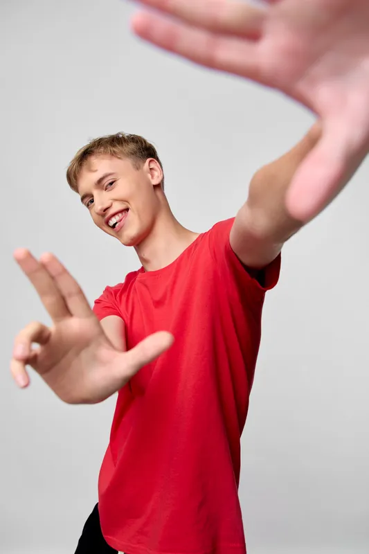 Dynamic movement of a smiling young man in a red shirt performing energetic gesture in a studio, capturing motion and interaction with the frame for active lifestyle, dance, fitness or performance