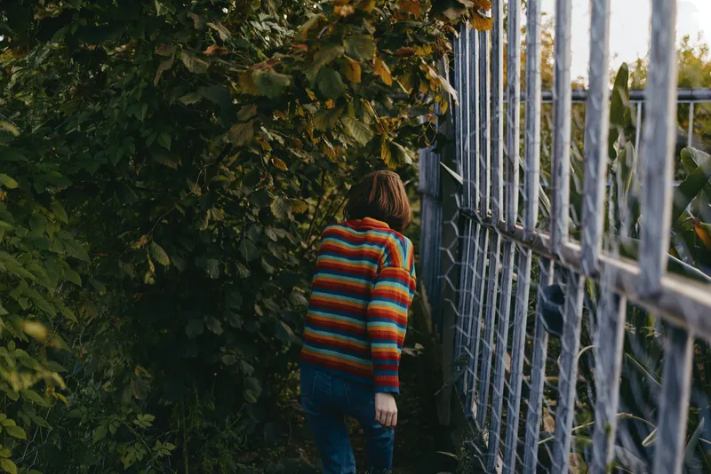 Young adult woman in a striped sweater walking along a metal fence through dense foliage, casual outfit and jeans, back view leaning forward in curious exploration outdoors.