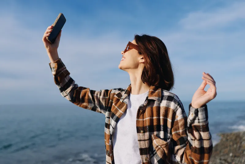 Woman by the sea in a plaid shirt and sunglasses takes a selfie with a phone, basking in bright sun. Ocean view, clear sky, and a relaxed, joyful outdoor moment.