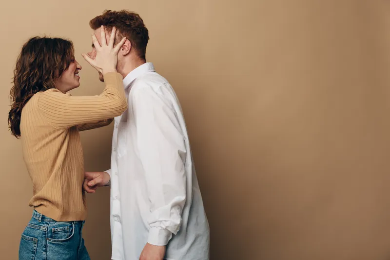 Young woman playfully covers young man face with hands while smiling, standing close on beige background. Concept of fun, relationship, interaction, and casual lifestyle between couple.