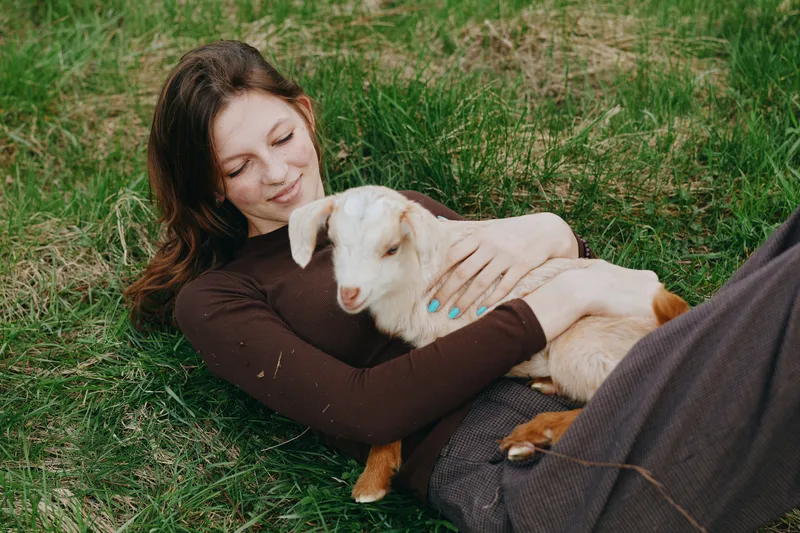 Relaxed woman lying on grass, cuddling a goat kid in a peaceful outdoor scene, casual clothing, soft light, close bond, simple joy, rural lifestyle, warm atmosphere, gentle affection, nature inspired