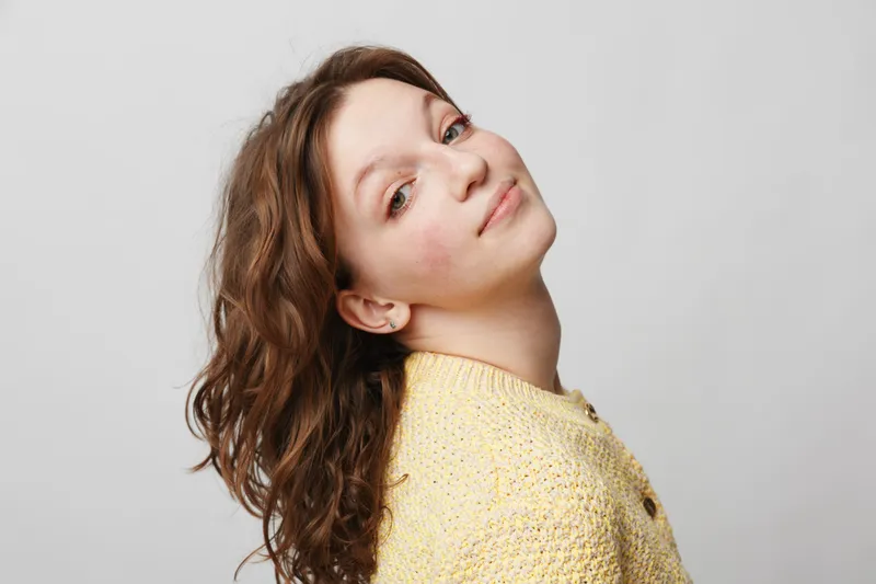 Young woman with curly brown hair wearing yellow sweater looks over shoulder with soft smile on light grey background. Concept of natural beauty, confidence, and casual portrait.