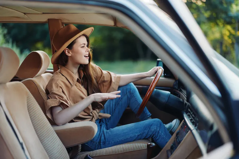 Happy young woman driving an antique car, wearing a stylish hat and casual outfit, enjoying a sunny day outdoors, embodying freedom and adventure.