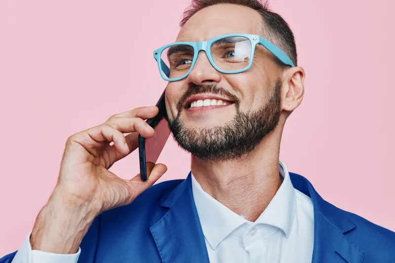 Professional man in a blue suit talks on the phone against a solid pink background, displaying confidence, focus, and optimistic energy.
