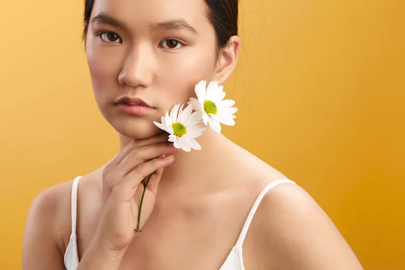 asian woman holds delicate daisies near her chin against a warm orange studio backdrop, soft lighting highlights clear skin, calm expression, and a gentle floral portrait.