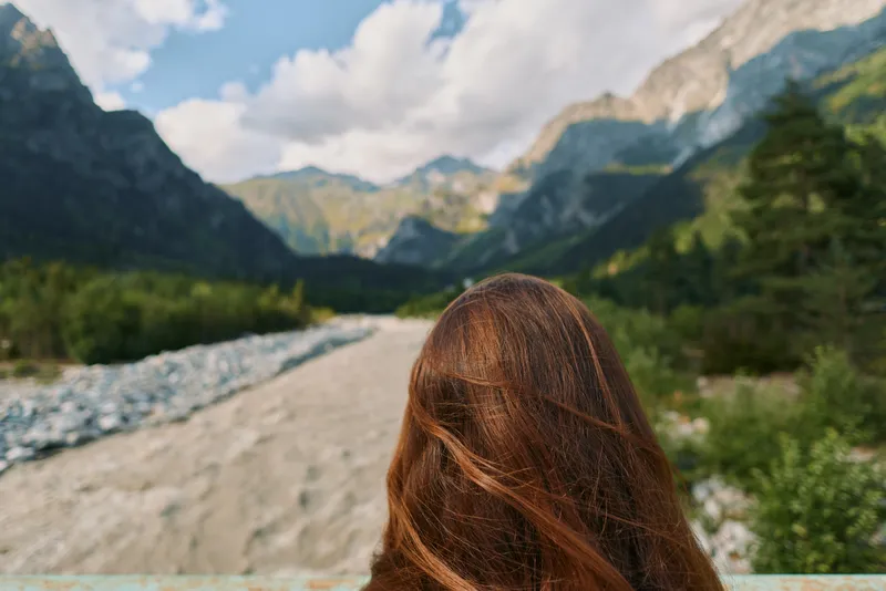 A person with long hair stands outdoors in a rugged mountainous landscape, embracing nature and a calm breeze, creating a peaceful, scenic moment.