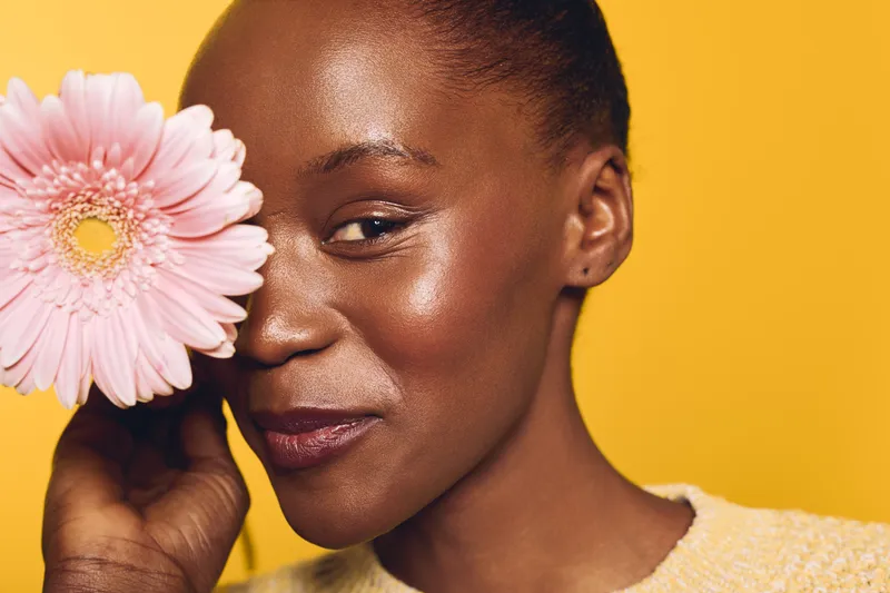 Close-up of smiling dark skinned African woman holding pink flower near face on yellow background. Concept of natural beauty, skincare, floral, and positive mood of black female model.
