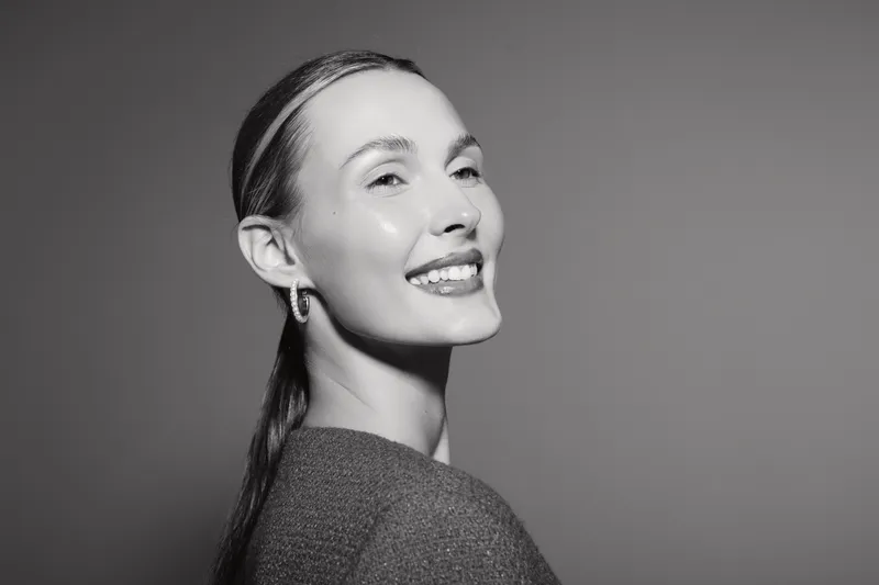 Smiling white woman with light skin and slicked back hair poses against grey background. Portrait in black and white.