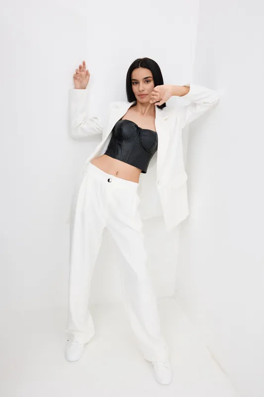 Woman posing confidently in white pantsuit and black corset top against bright minimalist background, showcasing modern fashion and stylish elegance.