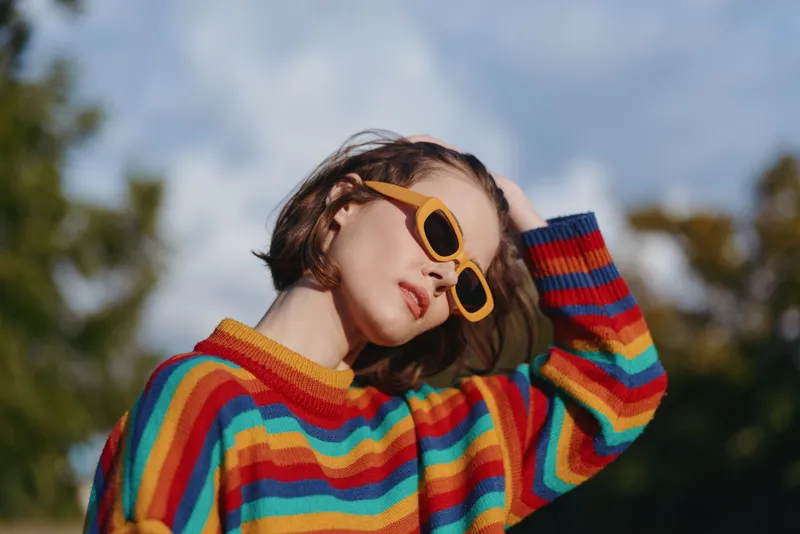 Woman in a striped sweater and yellow sunglasses, short hair styling in an outdoor portrait; young woman leaning back, casual outfit and relaxed thoughtful pose under blue sky.