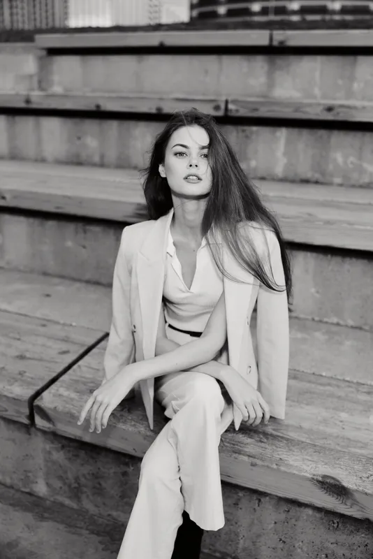 Confident woman in business attire sitting on wooden stairs, exuding elegance and professionalism in a monochrome setting.