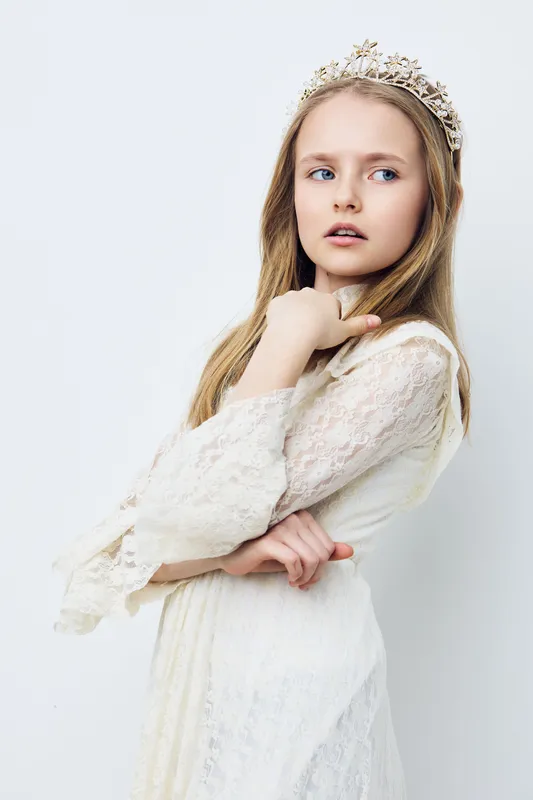 Young girl in a delicate white dress and crown, showcasing elegance, innocence, and beauty against a soft white background for children s fashion photography