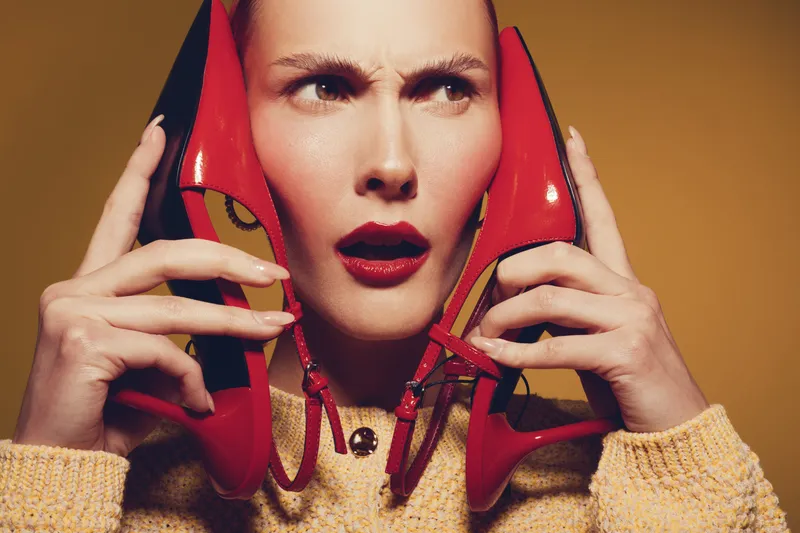 Confused woman holds red high heels near face, wearing yellow sweater. Studio portrait on brown background. Concept of fashion dilemma, style choice, and footwear emotions.