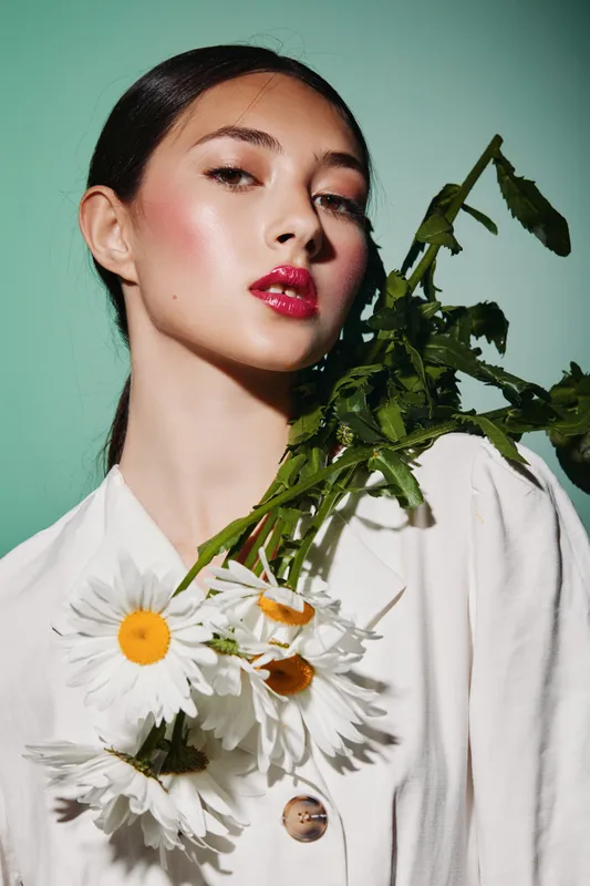 Young woman with bold red lipstick holds daisies against a mint green background wearing a simple white garment. Concept of beauty, fashion, and creative floral styling.