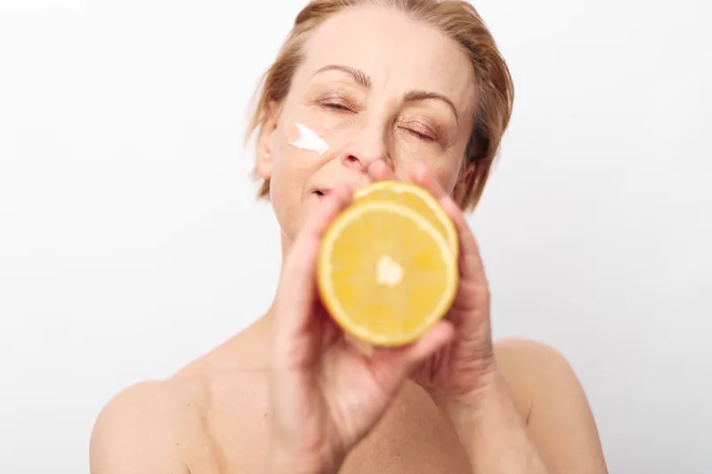 Mature woman with natural skin holds orange slice near face with moisturizer cream on cheek. Wellness concept for skincare, healthy lifestyle, and natural beauty on white background.
