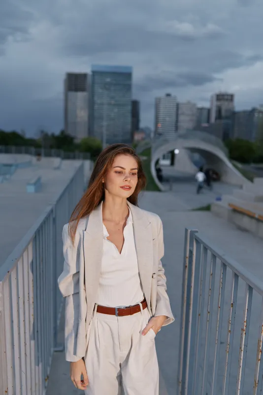 Confident businesswoman in stylish attire standing against a modern city skyline, showcasing professionalism and elegance during a cloudy day.