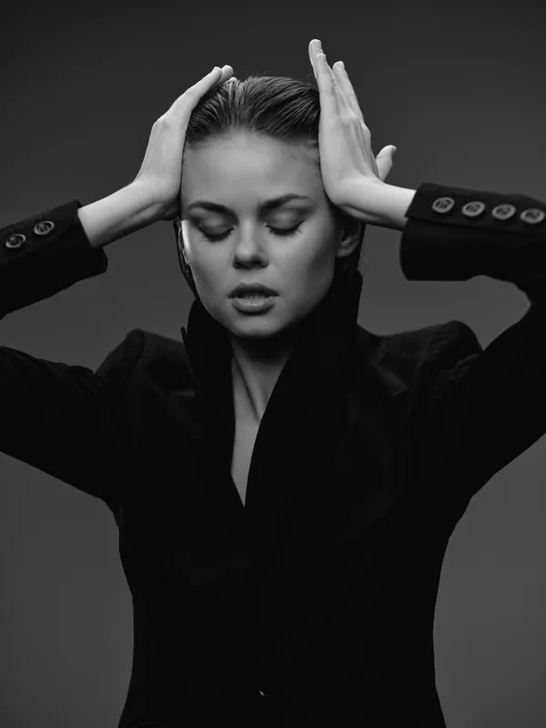 Black and white portrait of a woman in a black jacket with closed eyes and hands on her head expressing emotion