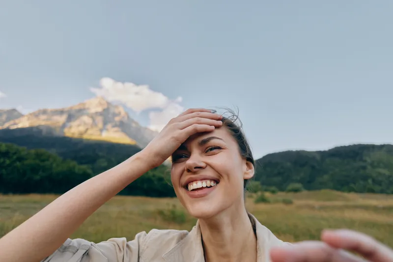 A happy young woman in casual clothes smiles outdoors with a mountain and forest in the background under a clear sky. Concept for lifestyle, nature, and joy.