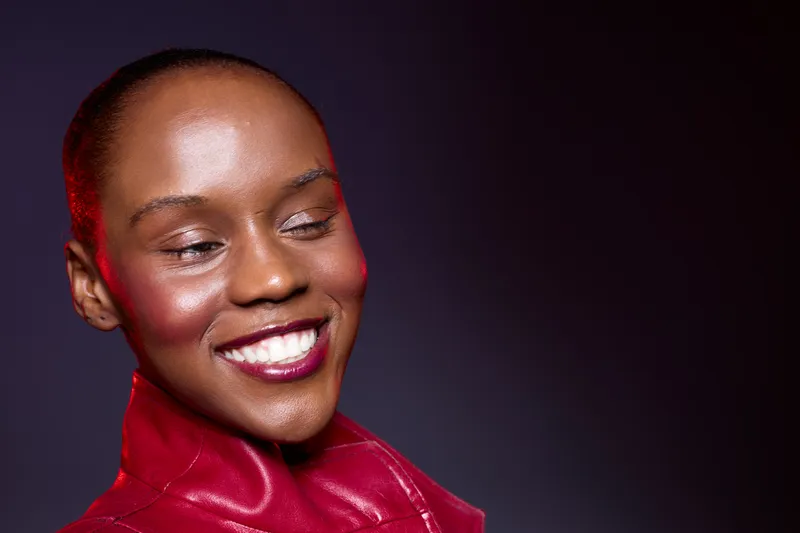 Close-up of a smiling African woman with dark skin wearing a red leather jacket. She looks down with closed eyes, expressing joy and confidence on a dark studio background.