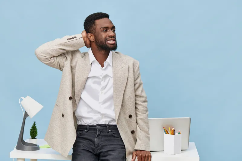 Confident man standing in bright office space with light blue background, wearing a beige blazer and white shirt, looking away with a relaxed expression, gesturing with hand near head, clean modern wo