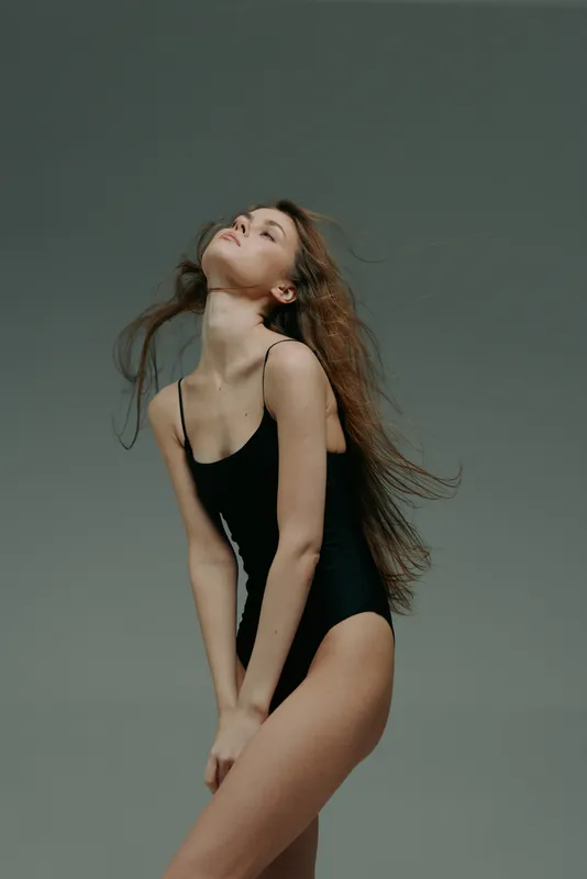graceful woman in black bodysuit posing with closed eyes and flowing hair against neutral gray background