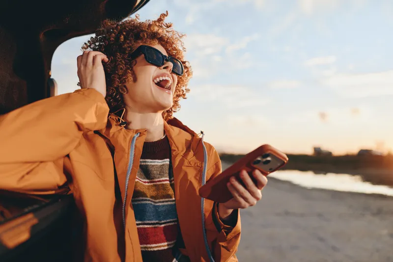 Smiling woman with curly hair wearing sunglasses, holding a smartphone at the beach, outdoors lifestyle moment. Orange jacket and warm sunset light create a joyful travel relaxed mood.