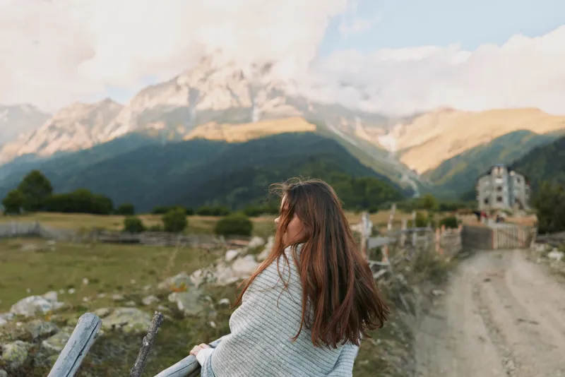 A woman enjoys a calm lifestyle moment on a hillside, gazing at mountains, embracing outdoor scenery, soft natural light, peaceful mood, candid travel vibe