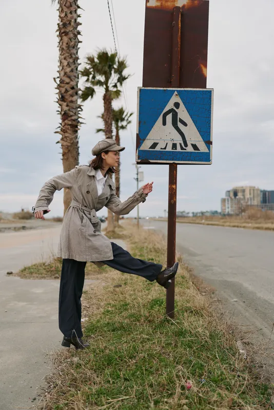 Woman in trench coat and cap stretches leg near pedestrian crossing sign on deserted road with palm trees. Concept of urban lifestyle, exercise, and casual fashion outdoors.