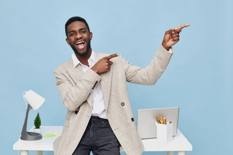 Smiling man with dark skin, wearing a beige sweater, pointing to the side while standing in a bright modern office with a desk, laptop, and decorative plant against a blue background. People lifestyle
