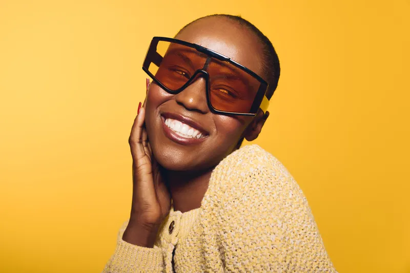 Happy smiling black woman with dark skin wearing oversized sunglasses and a beige sweater poses against yellow background. Concept for fashion, beauty, confidence, and stylish accessories.