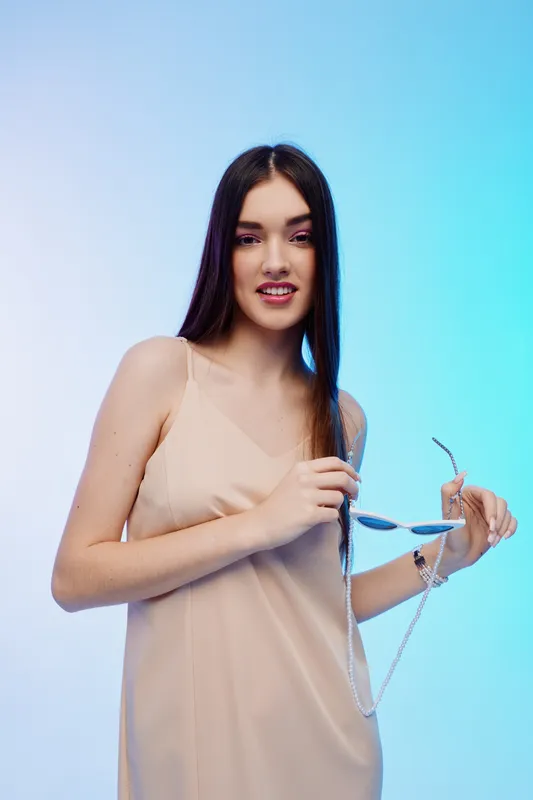 Young woman with long dark hair in a beige dress holding sunglasses against blue gradient background. Concept of beauty, fashion, and summer style with confident expression.