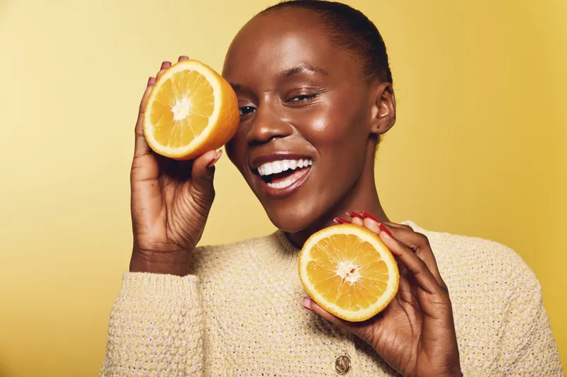 Smiling African woman with dark skin holds two orange halves near face against yellow background. Concept of healthy lifestyle, vitamin C, wellness, and natural beauty with citrus fruit.