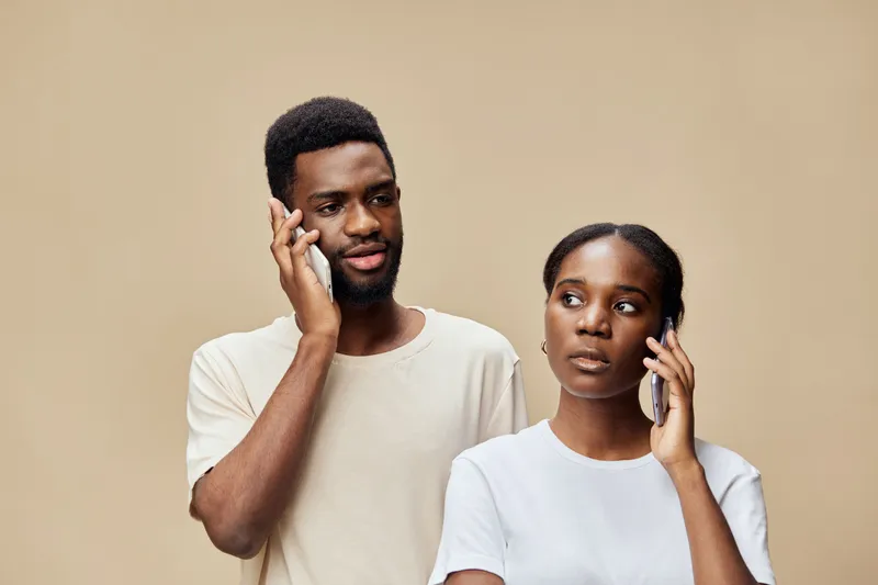 A young African couple engaged in a serious discussion on mobile phones against a neutral beige background, expressing different emotions, featuring casual attire