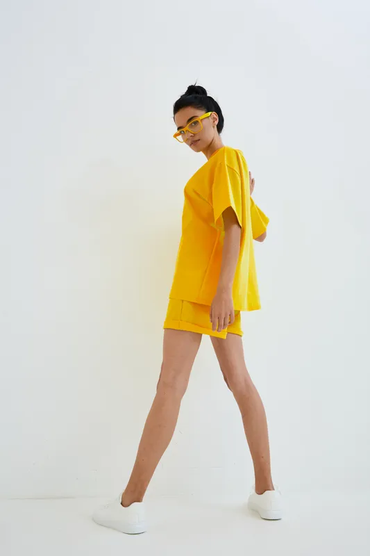 Slim woman in bright yellow casual outfit and glasses posing confidently on white background, showcasing modern street style and vibrant summer fashion.