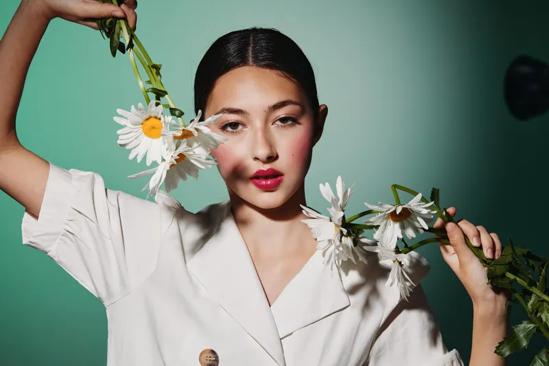 Fashion model with smooth skin holds fresh daisies close to her face wearing elegant white blazer on teal background. Concept of beauty, nature and creative fashion portrait.