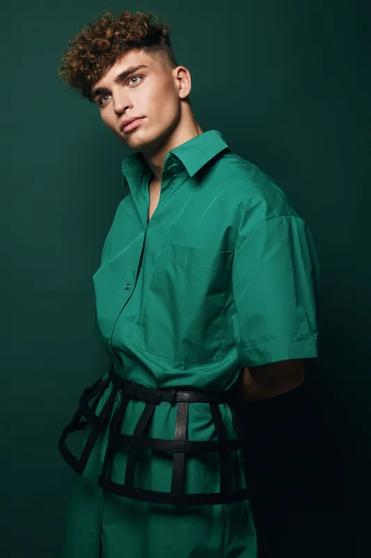 Young man with curly hair wearing green shirt and unique black belt posing against dark green background in studio portrait with serious expression and stylish look.