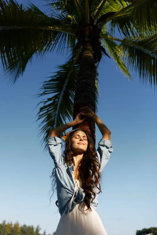 Relaxed woman standing under a tall palm tree, enjoying sunny weather with closed eyes, casual denim shirt, and long dark hair in a clear blue sky background.