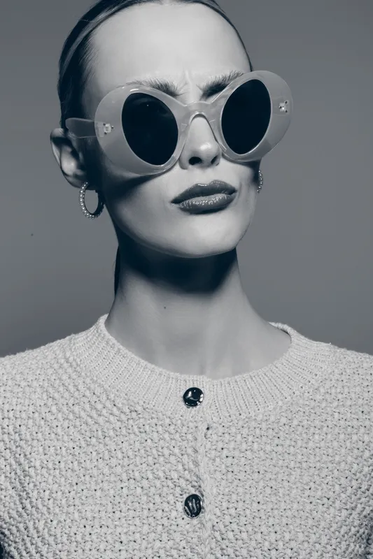 Stylish caucasian woman with pale skin wearing large round sunglasses and hoop earrings looks skeptical in black and white portrait.