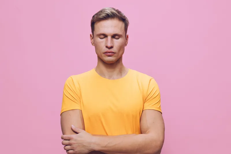 Confident young man with closed eyes, wearing bright yellow t-shirt, stands with arms crossed against pink background. Close-up studio portrait, casual style, facial expression shows calmness and self