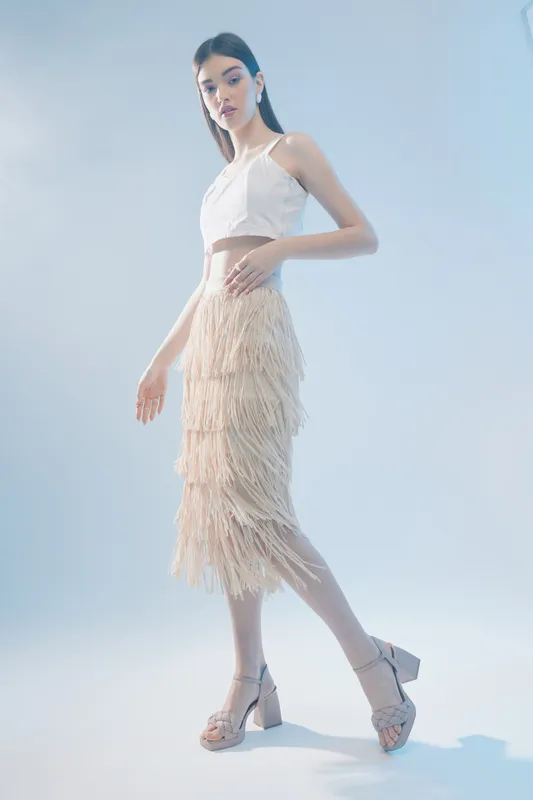 Slim woman beauty fashion model poses in a white crop top and fringed skirt against a light background. Studio shot with soft lighting highlights elegance and style.
