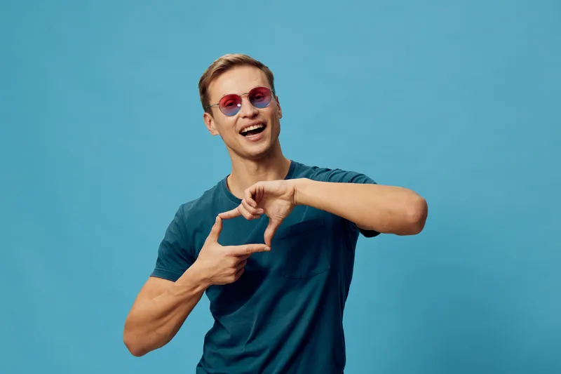 Joyful smiling young man with sunglasses wearing casual blue t-shirt expressing happiness and friendship isolated on blue background in studio concept. Lifestyle, positive emotion, fun, youth, portrai