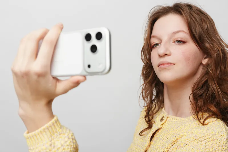 Young woman with curly hair wearing yellow sweater takes selfie with white smartphone on plain background. Concept of social media, technology, and casual lifestyle portrait.