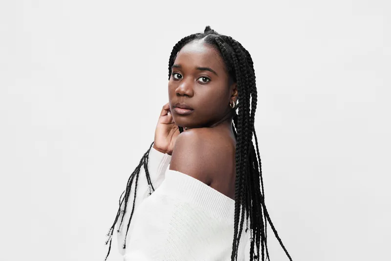 Young Black woman with braids in a white off shoulder sweater, showcasing confidence and elegance against a minimalist background