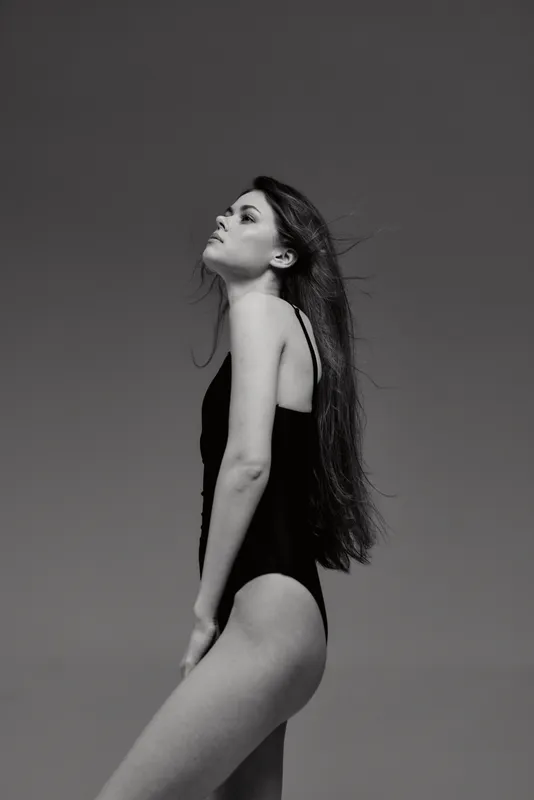 Black and white image of a woman in a black swimsuit posing thoughtfully against a plain background with long flowing hair.