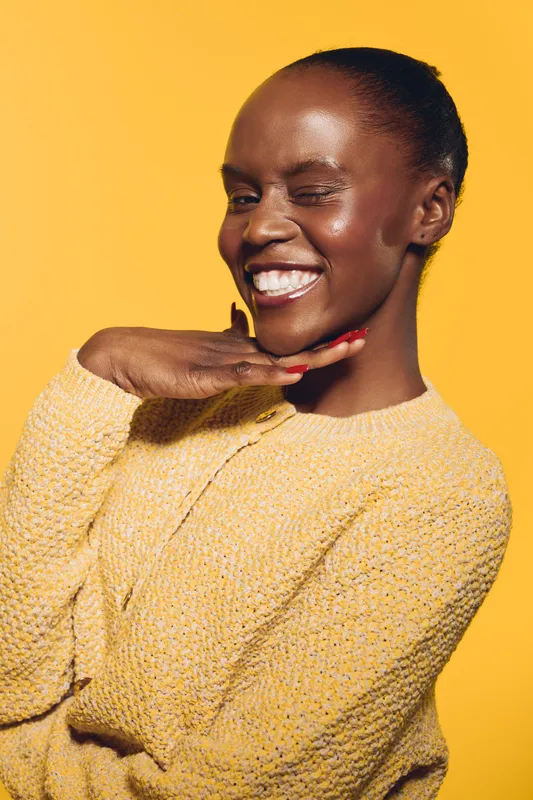 Happy African woman with dark skin winks and smiles wearing yellow sweater. Portrait on bright yellow background. Concept of joy, confidence, beauty, and positive emotions.